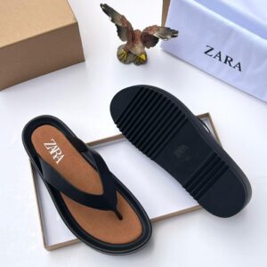 A pair of black T-strap flip flops with brown insole, placed on a white surface with a shoe box and bag in the background.