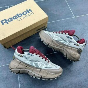 A pair of white and maroon Reebok Classic sneakers with tan soles, accompanied by a brown shoe box, exuding luxury and style.