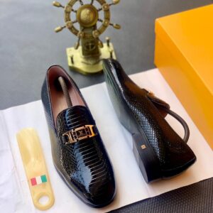 A pair of luxury black patent leather loafers with gold buckle, displayed on a white paper with a shoe horn and a golden ship wheel in the background, epitomizing high-class style and luxury.