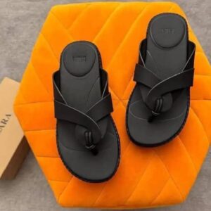 A pair of luxury black two-strap flat sandals on an orange hexagonal pillow, with a Zara box in the background, showcasing high-class products for the elite at Levonteluxe in Nigeria.