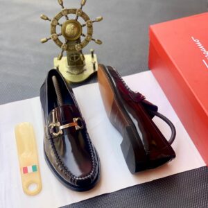 A pair of luxurious dark brown loafers with a gold buckle, placed on a white paper with a shoe horn and a red shoe box in the background, embodying high-class style and sophistication.
