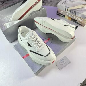 A pair of white Prada sneakers with red soles, displayed on top of their gray shoe box with a Prada tag