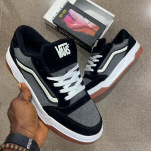 A pair of black and grey Vans shoes with white laces and brown soles, held by a hand with a watch and bracelet, showcasing the luxury and high-class design.