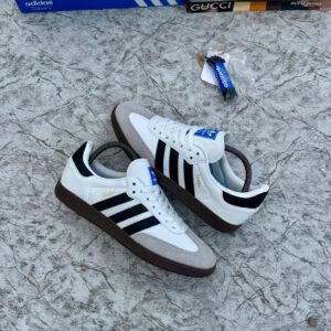 A pair of white and black Adidas Samba sneakers with brown soles, displayed on a marble surface with a blue Adidas box and luxury brand shoe boxes in the background.
