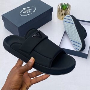 A hand holding a black Prada slide with the shoe box and sole in the background, showcasing luxury and high-quality craftsmanship.