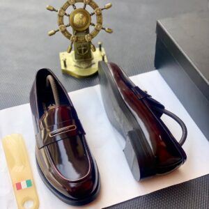 A pair of luxurious black leather loafers displayed on a white paper with a shoe horn and a decorative ship's wheel in the background, symbolizing high-end fashion and sophistication.