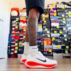 A close-up shot of a man's feet wearing white Nike ZoomX shoes with a black swoosh and red sole, standing in front of a collection of shoe boxes.