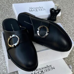 A pair of black leather mule shoes with a gold buckle, placed on top of an Alexander McQueen box, showcasing luxury and high-class design.