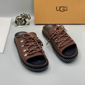 Luxury black UGG sandals with metal buckles and laces on a white surface, accompanied by a UGG box and a silver elephant statue, epitomizing high-class style and elegance.