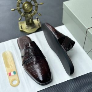 A pair of sleek black leather half shoes with a crocodile pattern, accompanied by a white shoe box and a decorative gold ship's wheel on a dark gray background.