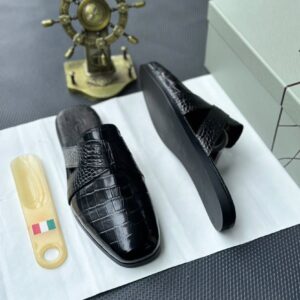 A pair of sleek black leather half shoes with a crocodile pattern, accompanied by a white shoe box and a decorative gold ship's wheel on a dark gray background.