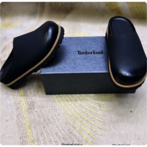 A pair of luxurious black Timberland half shoes with tan accents, resting on a gray box and a beige fabric background.