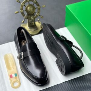 A pair of luxurious black leather loafers with a silver buckle, presented on a white paper with a green box and golden ship's wheel in the background, epitomizing high-end fashion.